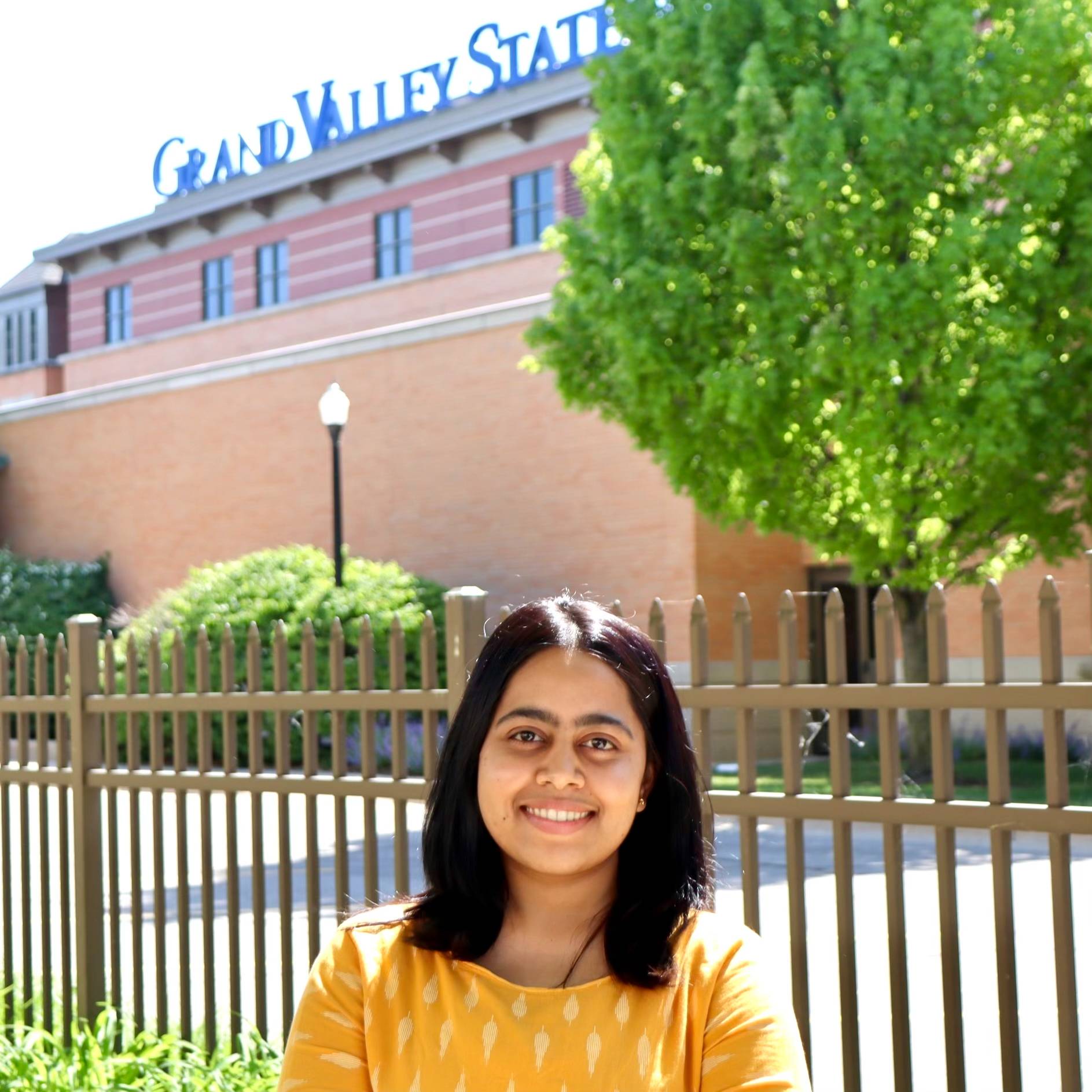 College of Health Professions Student Services student worker, Sunanda, smiling, stands in front of a Grand Valley building in the summer with a yellow shirt.
